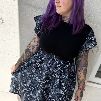 Vestido preto e branco com padrão complexo, parte superior preta, cinto preto, usado por pessoa com cabelo roxo e tatuagens, parede clara ao fundo.