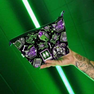 Necessaire preta com desenhos assustadores em roxo e verde, segurada por mão tatuada, fundo verde com luzes neon.