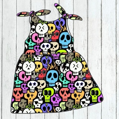 Vestido criança com padrão colorido de caveiras e tiras com laços em fundo de madeira clara.