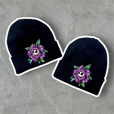 Duas gorros pretos com bordado de flor roxa e rosto de caveira no centro sobre fundo cinzento