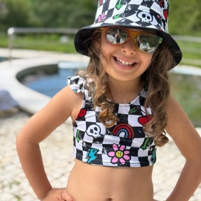 Criança com roupa de padrão quadriculado colorido e chapéu, junto a área externa com piscina.