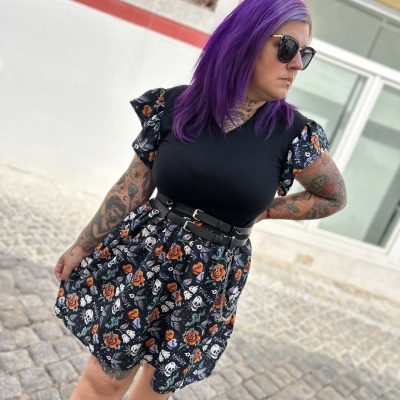 Vestido preto com padrão de caveiras e rosas, cinto preto, usado por mulher com cabelo roxo e óculos
