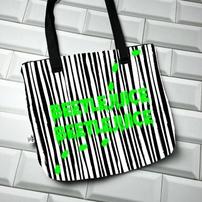 Mala tote bag riscas pretas e brancas com texto verde BEETLEJUICE sobre azulejos brancos