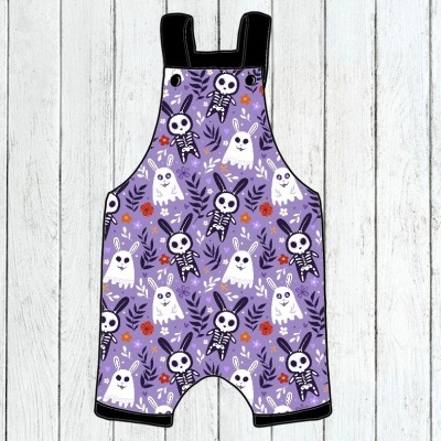 Macacão infantil roxo com padrão de Halloween de fantasmas e esqueletos em fundo de madeira branca.