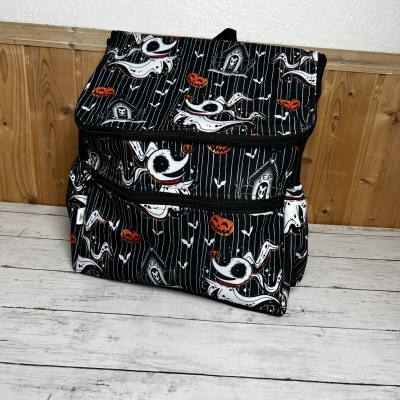 Mochila preta com padrão de fantasmas, abóboras e túmulos em ambiente interior com fundo de madeira e parede branca