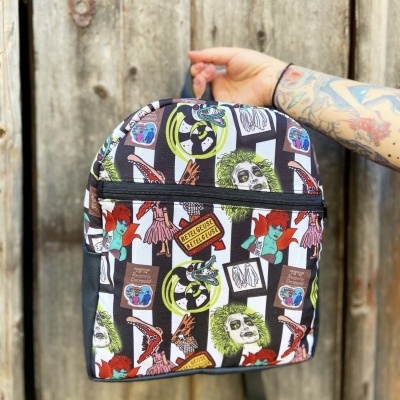 Mochila colorida com padrão de personagens e logótipos do filme Beetlejuice segurada por mão tatuada