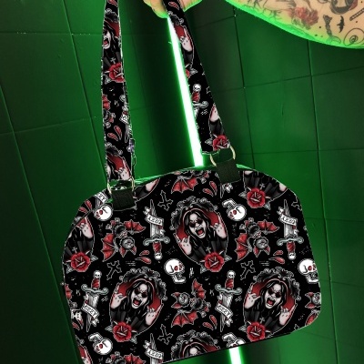 Mala de mão preta com padrão de caveiras, rosas e rostos femininos com uma mão tatuada segurando-a