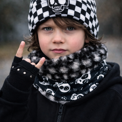 Criança com gorro quadriculado preto e branco com caveira, cachecol peludo cinza e preto, luva preta com tachas e casaco preto com fecho.