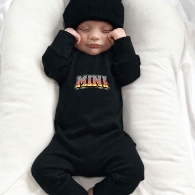 Bebé com macacão preto e gorro preto deitado numa almofada branca, com a palavra MINI no macacão.