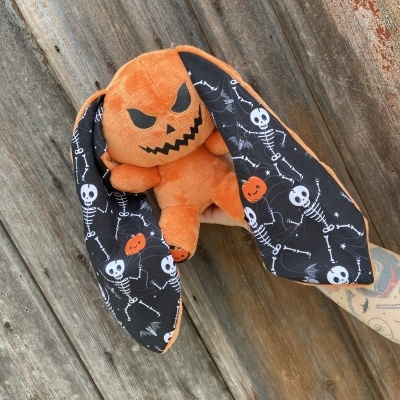 Boneco de peluche laranja com cabeça em forma de abóbora e orelhas longas com padrões de esqueletos