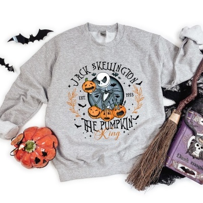 Sweatshirt cinzenta com estampado de personagem e texto Halloween, rodeada de adereços festivos