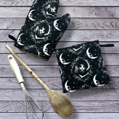 Conjunto de luva de forno e pega pretos com padrão lunar e rosto feminino ao lado de utensílios de cozinha sobre madeira cinzenta