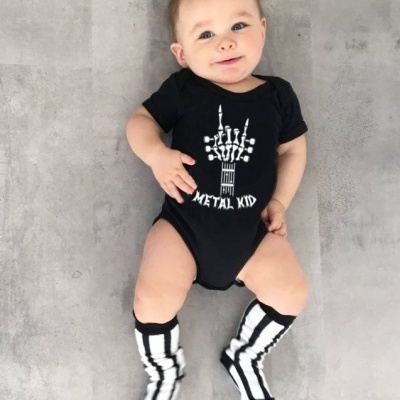 Bebé com body preto METAL KID e meias brancas e pretas deitadas no chão cinzento.