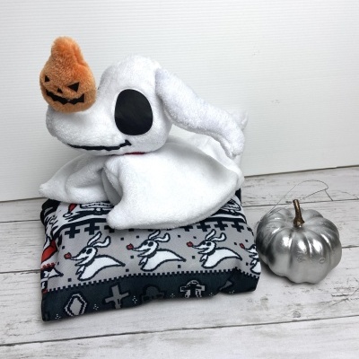 Peluche fantasma branco com abóbora laranja sobre manta com padrão de coelhos e abóbora prateada ao lado