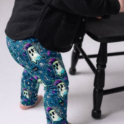 Calças azuis com padrão de fantasmas coloridos e casaco preto numa criança apoiada numa cadeira preta.