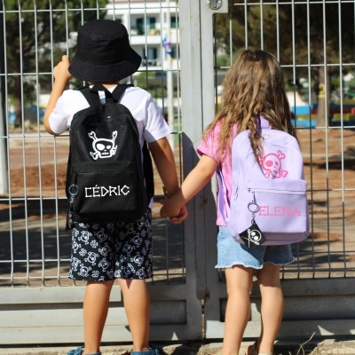 Crianças de costas com mochilas personalizadas, diante de grade metálica.