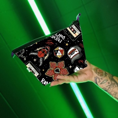 Bolsa triangular preta com padrão colorido e texto vários, segurada por mão tatuada com fundo verde iluminado