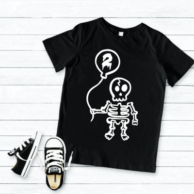 T-shirt preta com estampa de esqueleto e balão, ténis infantis pretos e brancos sobre chão de madeira clara
