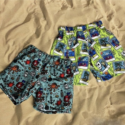 Dois calções de banho infantis com padrões coloridos sobre areia.