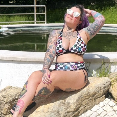 Mulher sentada numa pedra junto a piscina com biquíni xadrez colorido e várias tatuagens