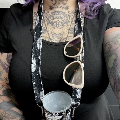 Mulher com tatuagens, vestido preto, segura copo com alça preta e óculos de sol bege