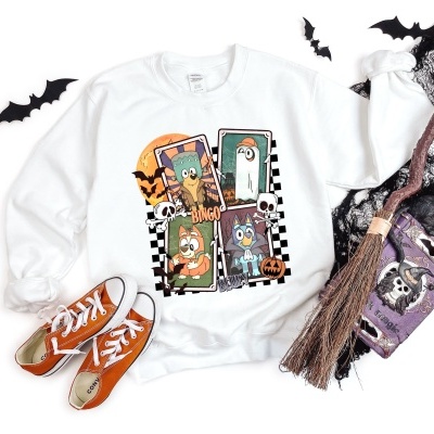 Sweatshirt branca com personagens animados e motivos de Halloween, sapatos laranja, vassoura e sacola roxa com caveira