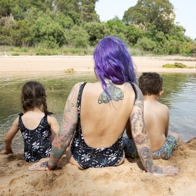 Mulher e duas crianças sentadas na areia junto a uma lagoa, fatos de banho com padrões florais e calções de banho coloridos