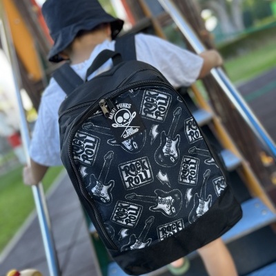 Mochila preta com padrão branco de guitarras e texto Rock & Roll vista nas costas de uma criança num parque infantil
