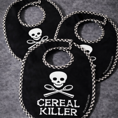 Três babetes pretos com padrão xadrez nas bordas e design de crânio e talheres cruzados com texto CEREAL KILLER