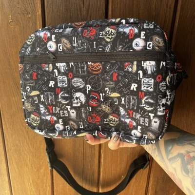 Bolsa tiracolo preta com padrão de caveiras, olhos e letras coloridas segurada à frente de porta de madeira.