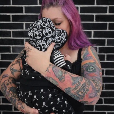 Mulher com cabelo rosa e tatuagens abraça bebé com roupa preta de caveiras