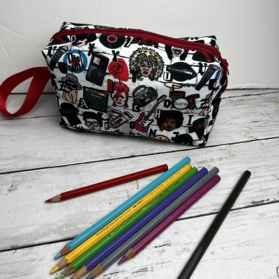 Necessaire branca com padrão colorido de desenhos musicais e fecho vermelho, junto a lápis de cor sobre mesa branca
