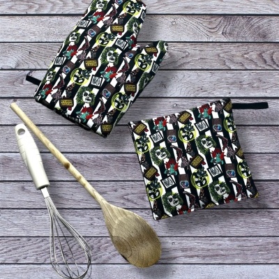 Conjunto de luvas de forno e suporte para panelas com padrão colorido sobre superfície de madeira clara, junto com utensílios de cozinha.