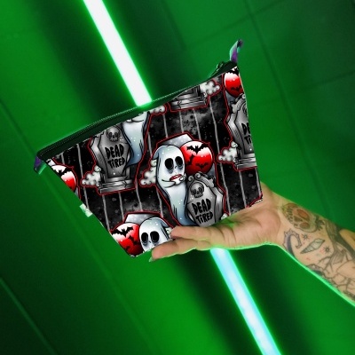 Necessaire preta com padrão de fantasmas e lápides, texto 'DEAD TIRED', segurada por mão tatuada