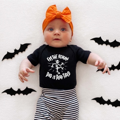 Bebé vestido com body preto estampado, calças de riscas e bandana laranja, com morcegos de decoração ao redor num fundo branco.