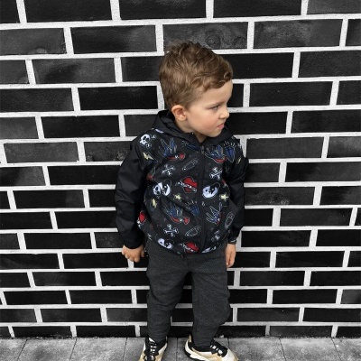 Criança com casaco preto estampado e calças cinza em frente a parede de tijolos pretos