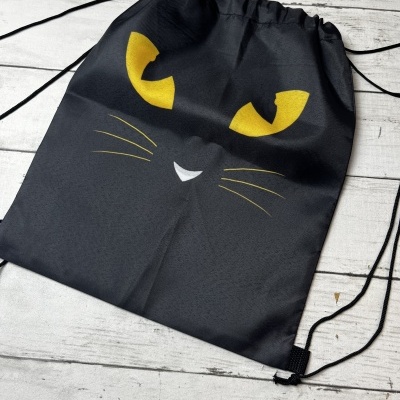 Mochila preta com rosto de gato em tecido, cordões pretos, sobre madeira clara.