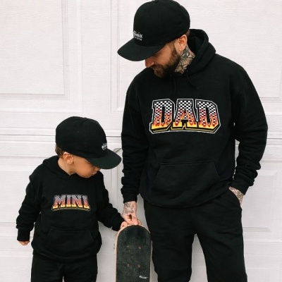 Duas pessoas vestem camisolas pretas com texto MINI e DAD, bonés pretos e seguram um skate preto em frente a uma porta branca.