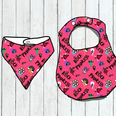 Babete e bandana rosa com padrão 'ROCK PRINCESS' e desenhos coloridos sobre fundo de madeira clara
