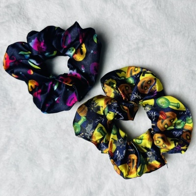 Dois scrunchies com padrão Halloween apresentando abóboras e morcegos, um com fundo preto e outro amarelo, sobre superfície branca.
