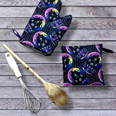 Luvas e pega-luvas de cozinha com padrão de casas amarelas e roxas sobre fundo listrado preto ao lado de utensílios de cozinha sobre madeira cinza