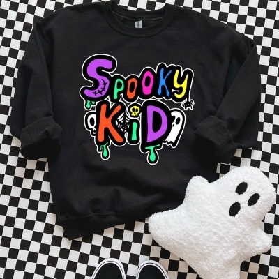 Sweatshirt preta com estampa Spooky Kid colorida sobre piso xadrez, peluche de fantasma branco e ténis pretos.