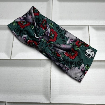 Faixa de cabelo com padrão de gatos, flores vermelhas, folhas verdes, texto 'I AM NOT CUTE' e caveira em azulejos brancos.