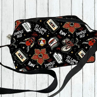 Mala preta com padrão colorido de flores, bicicletas, dados, fitas cassete, bonés e textos sobre fundo de madeira clara