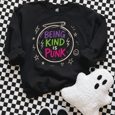Sweatshirt preta com frase colorida BEING KIND IS PUNK sobre fundo xadrez, perto de almofada fantasma e sapatos pretos com atacadores brancos.