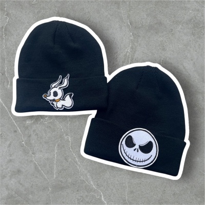 Dois gorros pretos com bordados distintos, um com pato e outro com cara sorridente, sobre superfície cinzenta.