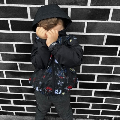 Criança com casaco preto com capuz e padrões coloridos em frente a parede de tijolos pretos.