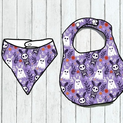 Babete e bandana roxos com padrão de esqueletos, fantasmas e folhas sobre fundo de madeira