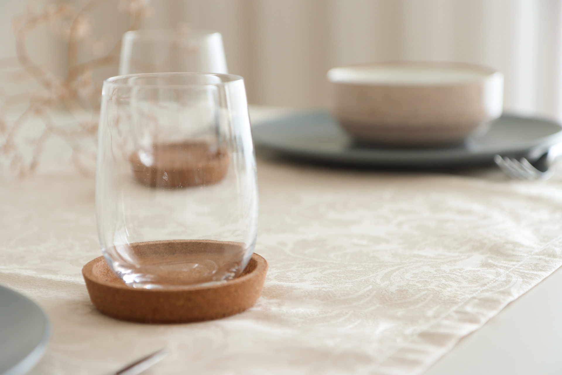Copos de vidro sobre bases de cortiça numa mesa com toalha bege e pratos cinza ao fundo