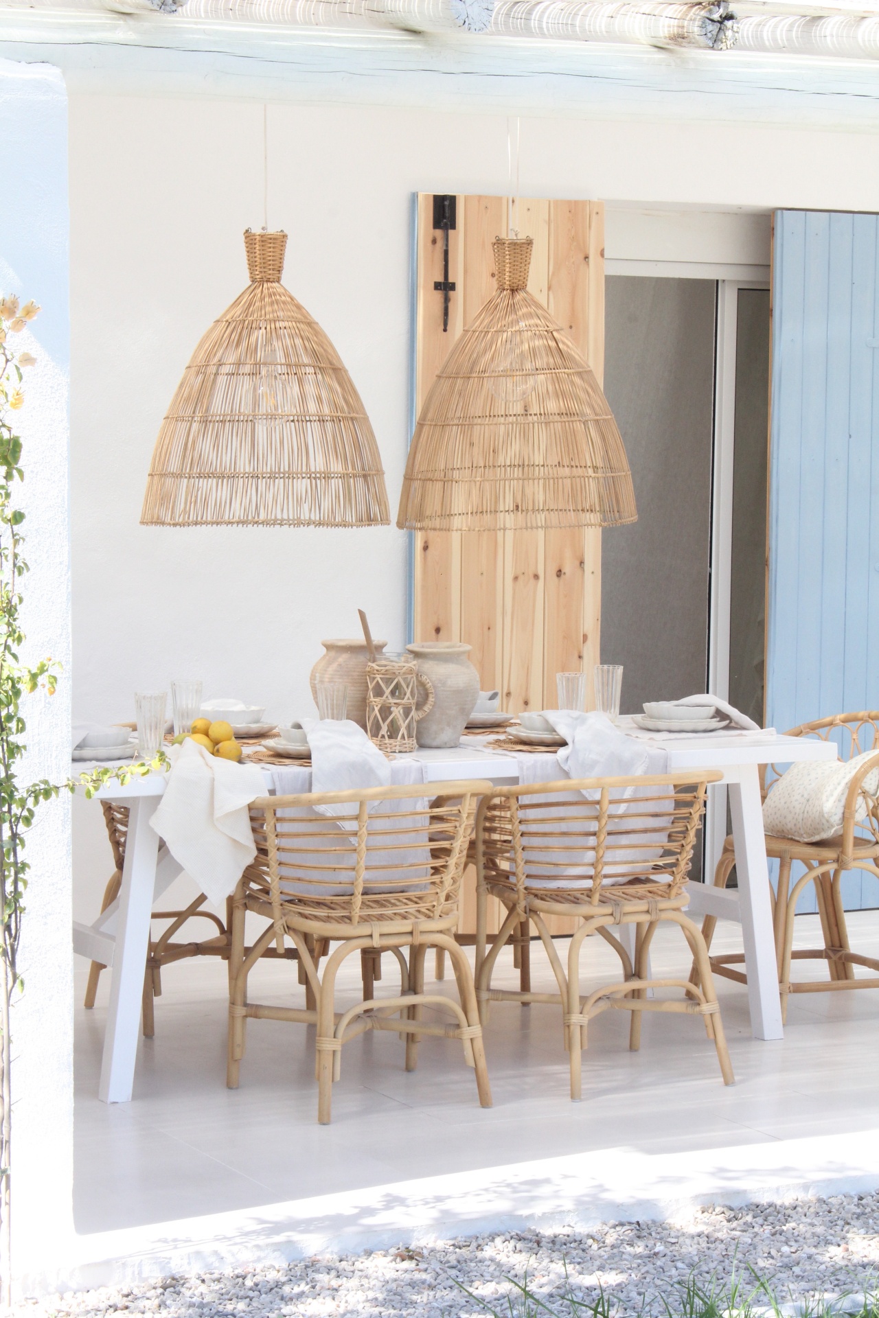 Conjunto de mesa de jantar branca com cadeiras de rattan, luminárias de vime pendentes, em ambiente claro e arejado.